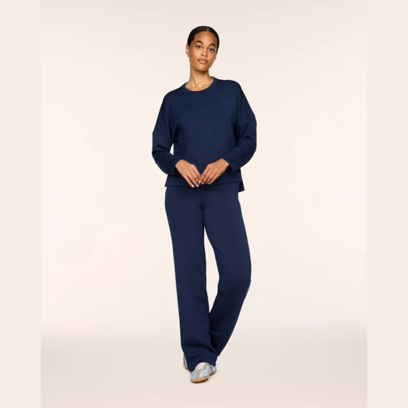 A woman modeling a matching navy blue lounge set consisting of a long-sleeve crewneck sweatshirt and wide-leg sweatpants, standing against a plain light beige studio background.