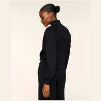 A model shown from the back wearing a black ribbed mock-neck long-sleeve top and matching black ribbed trousers against a solid off-white background.