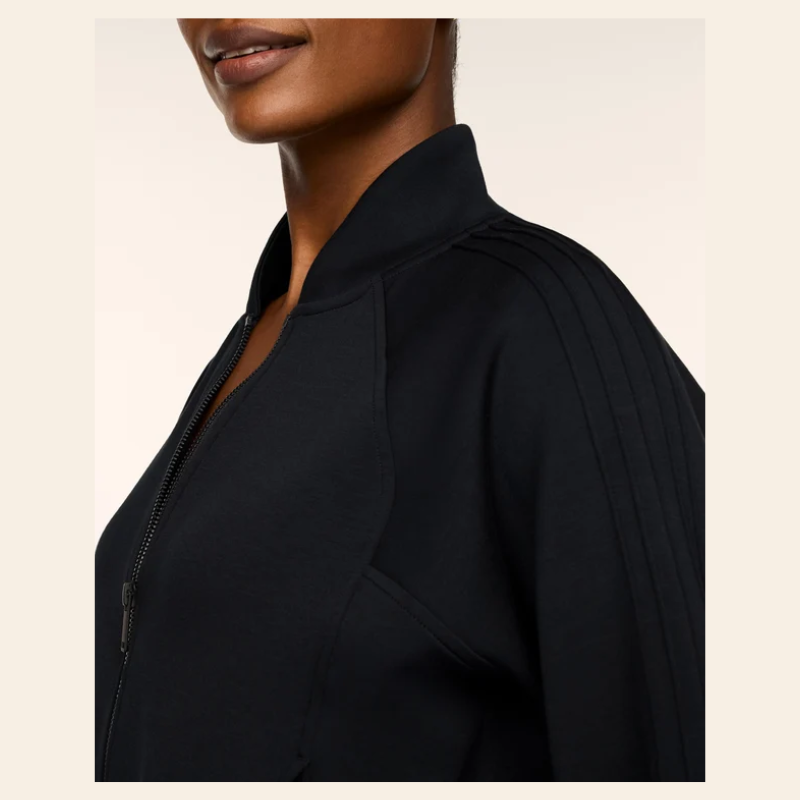 Close-up side view of a model wearing a black zip-front jacket with a structured collar and shoulder pleats, shown against a light beige background.