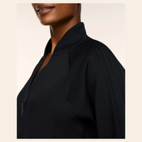 Close-up side view of a model wearing a black zip-front jacket with a structured collar and shoulder pleats, shown against a light beige background.