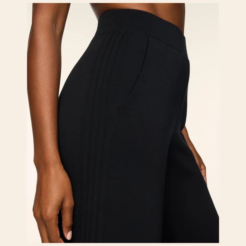 Close-up of a model wearing black pleated wide-leg trousers against a light beige background.