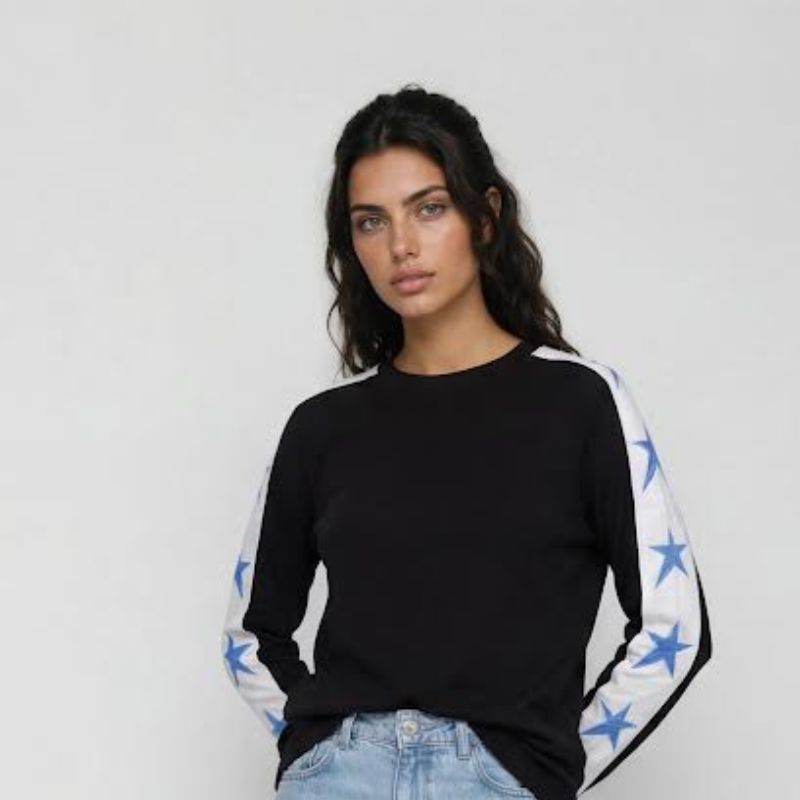 A woman modeling a black long-sleeve top with white sleeves featuring blue star patterns, paired with light blue jeans, against a plain light gray background.