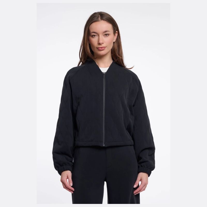 A woman modeling a black zip-up bomber jacket with a ribbed collar, cuffs, and hem, paired with black pants against a light gray studio background.