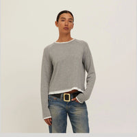 A model wearing a light gray long-sleeve raglan sweater with white ribbed trim at the crew neck, cuffs, and hem, paired with blue jeans and a black belt, against a plain off-white background.