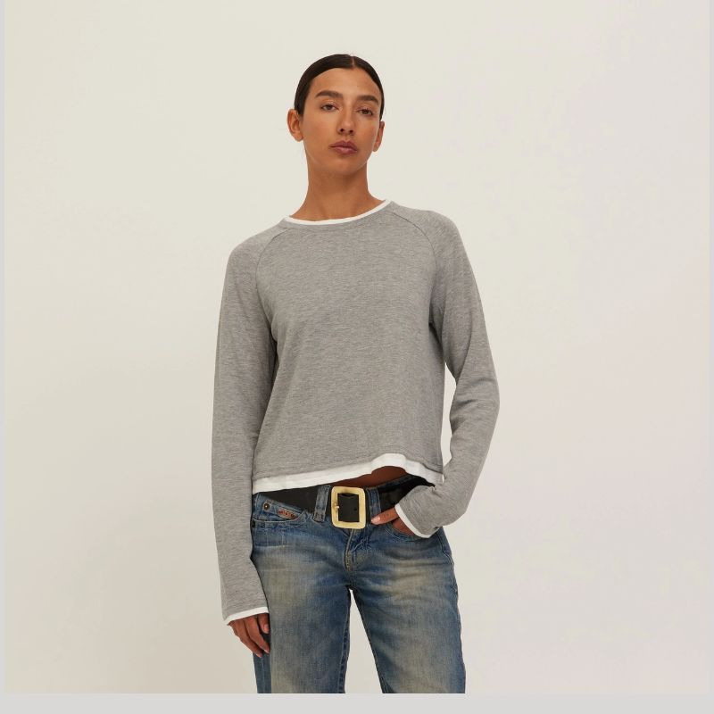 A model wearing a light gray long-sleeve raglan sweater with white ribbed trim at the crew neck, cuffs, and hem, paired with blue jeans and a black belt, against a plain off-white background.