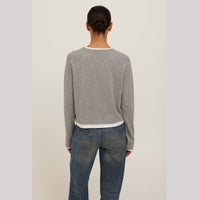 A person viewed from the back wearing a light gray cropped long-sleeve sweater with white ribbed trim and blue jeans, standing against a plain light gray studio background.