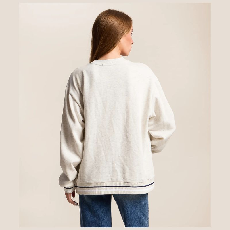 A woman viewed from the back wearing an oversized off-white crewneck sweatshirt with black stripe detailing at the hem and cuffs, paired with blue jeans, against a solid beige background.