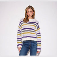 A woman modeling a fuzzy, long-sleeve knit sweater with horizontal stripes in purple, mustard yellow, gray, and white, paired with blue jeans against a plain white studio background.