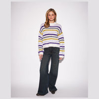 A woman modeling a fuzzy, long-sleeve knit sweater with horizontal stripes in purple, yellow, and white, paired with dark blue flared jeans, against a light gray studio background.