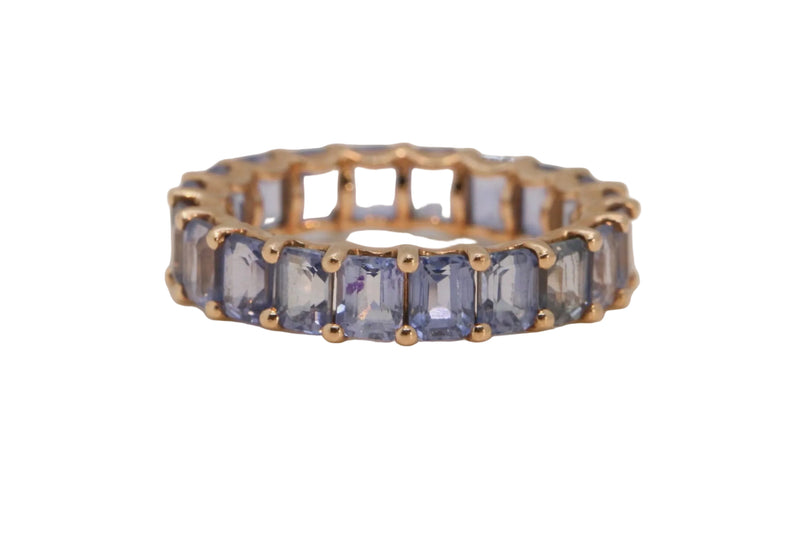 A gold eternity ring featuring a continuous row of rectangular blue gemstones in a prong setting, shown on a plain light background.